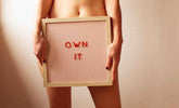 Own It framed sign held by a person, featuring red letters on a striped background.