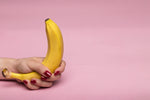 Banana held in a hand with red nails against a pink background.