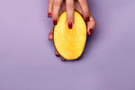 Fresh mango held in hand with red nails against a purple background.