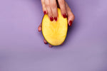 Mango slice held in hand with red nails against a purple background.