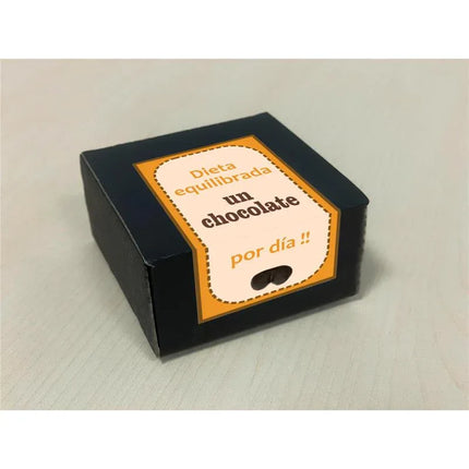 Black box with orange and white label featuring Spanish text "Dieta equilibrada un chocolate por día" on a light surface.