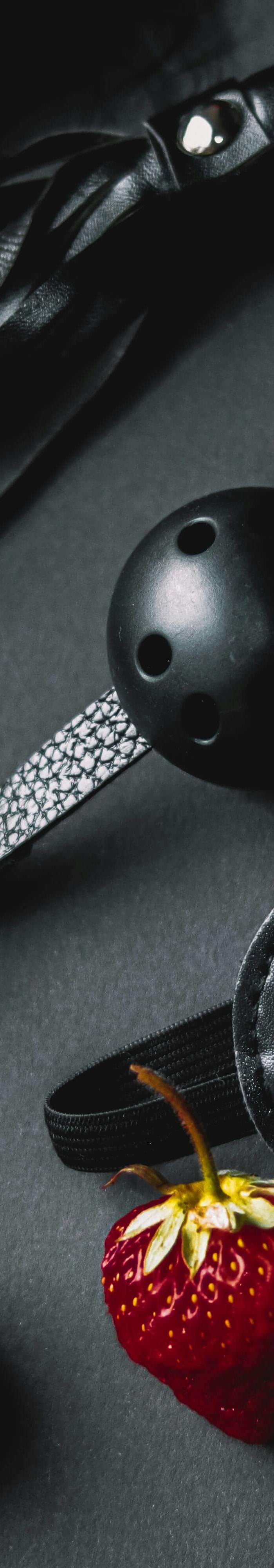 Black leather BDSM accessories with strawberries on black background, including whip, blindfold, and ball gag.