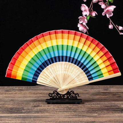 Wooden Fan LGBT with rainbow design, folding bamboo fan, lightweight and portable, displayed on a wooden table with pink blossoms.